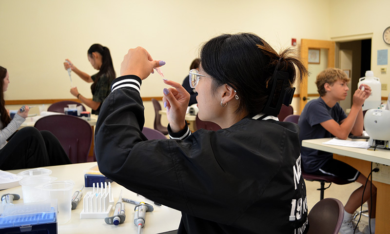 A student carefully examines a small liquid sample in a clear microcentrifuge tube and other students conducting experiments in a DNA science laboratory